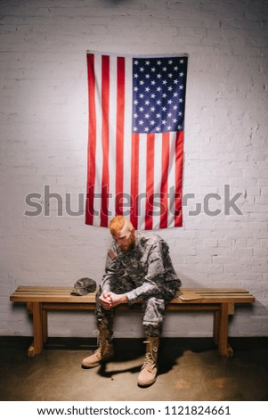 soldier in military uniform sitting on wooden bench with american flag on white brick wall behind, 4th july holiday concept