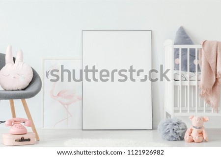 Flamingo poster standing on the floor behind empty mockup poster in white baby room interior with cute pillows and wooden crib. Place your photo here Royalty-Free Stock Photo #1121926982