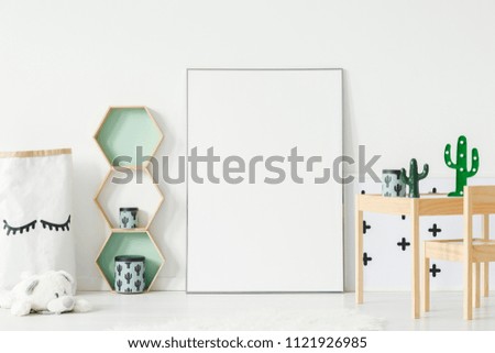 Wooden shelves with cactus boxes and decorations on small table standing in white kid room interior with mockup poster on the floor. Paste your photo here Royalty-Free Stock Photo #1121926985