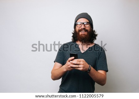 Portrait of a happy young bearded hipster man holding mobile phone while standing and looking at camera over white background Royalty-Free Stock Photo #1122005579