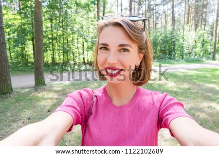 Young cute cheerful girl in green park at sunny daylight taking pictures of herself