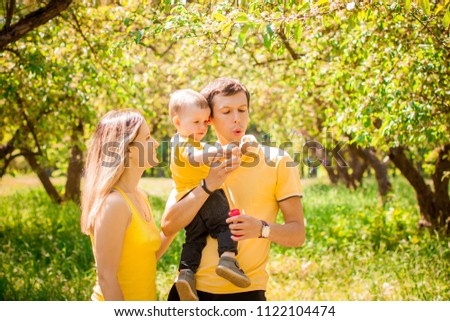 happy family with one child in yellow T-shirts sitting in the garden on the grass smiling and pointing a finger forward on blowing soap bubbles. Sunny summer mood, one little boy and dad with mom