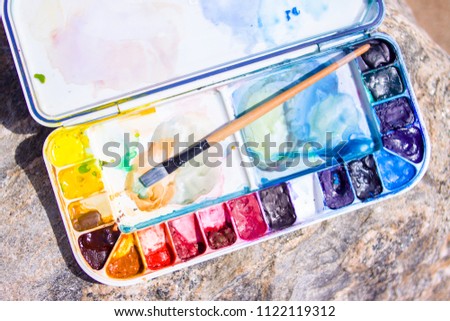 creation, drawing and freedom concept - Artistic equipment: paint brushes, palette and paintings on rock in nature at sunny summer day