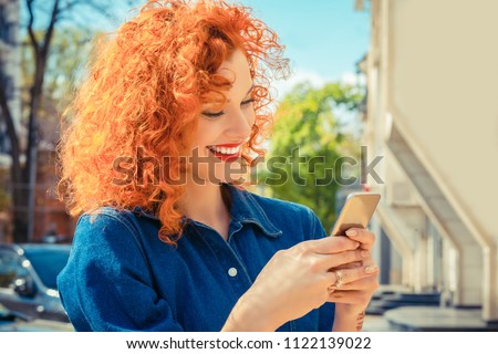 Young beautiful red curly hair woman, smiling, looking her mobile phone texting, reading sms message. Isolated city on background. Positive human emotions face expression feelings.