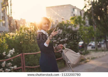 Girl holds backpack in her hands and turns her face in frame. She is walking along city street. Street fashion. Sunset sun in background.