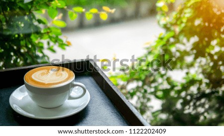 a cup of hot coffee with milk art on wooden black plate at the left of photo from side view beside the glass window see through outdoors with green view.