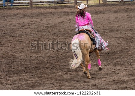 A cowgirl in a beautiful pink outfit rides on the back of her decorated blond colored horse. The rider has long black hair and wears a white cowboy hat. She and the horse are inside an arena with dirt