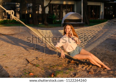 Young woman sitting in hammock and talking by smartphone, summer vacations. Concept of resting in tropicak resort and advantageous tariff plan, modern technology.