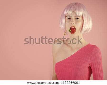 Female fashion concept. Girl in artificial hair hold red berry in mouth. Fruit and vitamin. Skincare, spa and health. Beauty and fashion. Woman in wig has no makeup on pink background, copy space