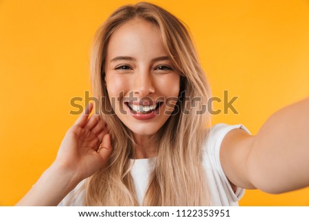 Cheerful young blonde girl taking a selfie with outsretched hand isolated over yellow background