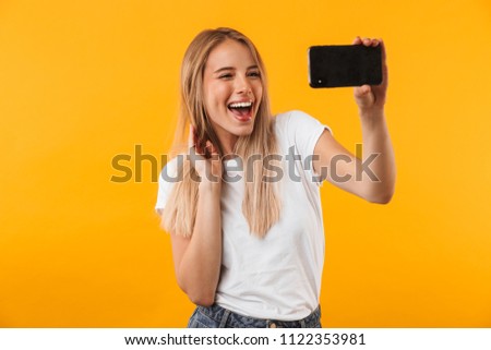 Cheerful young blonde girl taking a selfie with mobile phone isolated over yellow background