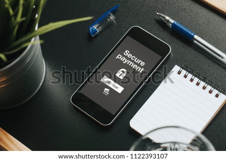 Diagonal view of mobile phone with payment service in the screen on a black table with a notebook, a pen, a glass of water and a green plant.