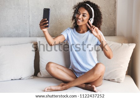 Smiling african woman in headphones taking selfie with mobile phone while sitting on a couch at home