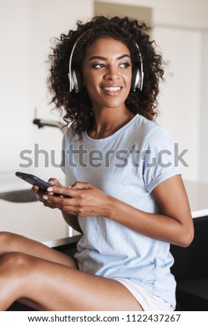 Attractive african woman in headphones using mobile phone while sitting on a chair at home