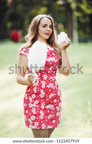 Handsome girl holding candyfloss in her hand and eating it. Adorable lady wearing flowery red dress and with chestnut hair posing with cotton candy. Background of city park.