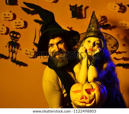 Little witch wearing black hat. Girl with cute face on yellow background with decor. Kid in spooky witches costume in yellow light holds carved jack o lantern. Halloween party and decorations concept
