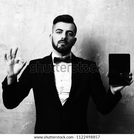 OK sign and black tablet in hands of bearded businessman wearing suit and red bow tie, on old painted wall background. Idea of technologies and best choice