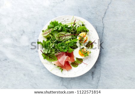 Mixed leaf salad with egg, serrano ham, and herbs. Healthy diet. Flat lay. Home made food. Paleo diet. Bright stone background. Top view. Copy space. 