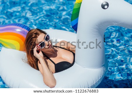 A young beautiful girl with a beautiful figure in a black swimsuit and sunglasses is resting and tanning in a swimming pool with an inflatable unicorn.