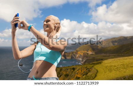 sport, technology and healthy lifestyle concept - smiling young woman with smartphone, earphones and fitness tracker listening to music over big sur hills and pacific ocean background in california