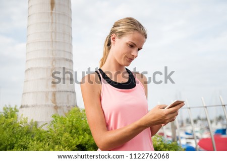 Young woman standing by a palm tree surrounded by green shrubs smiling as she looks at her cell phone