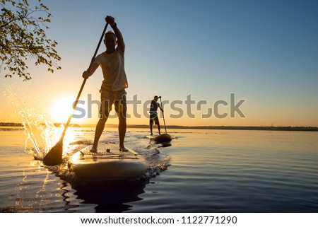 Men, friends sail on a SUP boards in a rays of rising sun. Stand up paddle boarding - awesome active recreation in nature. Backlight.  Royalty-Free Stock Photo #1122771290