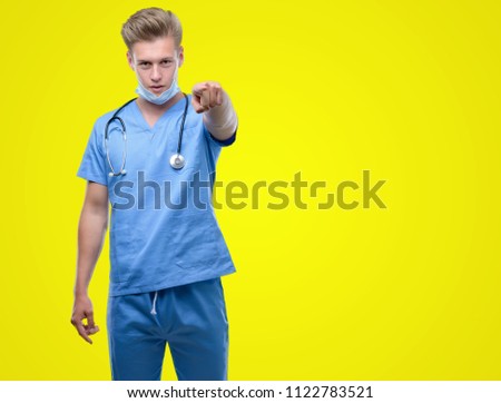 Young handsome blond doctor pointing with finger to the camera and to you, hand sign, positive and confident gesture from the front