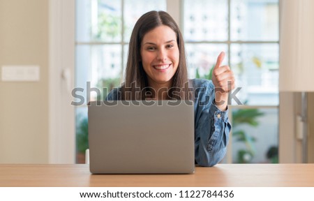 Young woman using laptop at home happy with big smile doing ok sign, thumb up with fingers, excellent sign