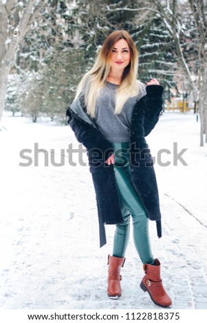 Young slim blonde woman wearing in grey sweater, emerald leggins, black fur  coat and brown stylish boots in winter city park. Girl rejoices in winter. Winter fir on the background. Full length photo.
