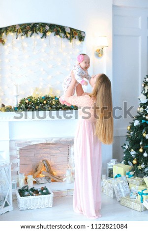 Young woman wearing pink dress and holding female child in arms and standing near decorated fireplace and Christmas tree. Concept of winter holidays and Christmas decorations.