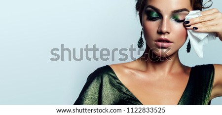 beautiful young girl with a bright make-up and in a shiny green dress striatet makeup from her face with a wet napkin. Hairstyle - curls are gathered in a bun.fashion, beauty, makeup, cosmetics. Royalty-Free Stock Photo #1122833525