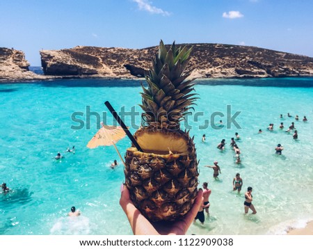 Close up of tropical pineapple cocktail against blue lagoon with turquoise water. Beach and beautiful sea. Travel concept. Pineapple fruit with margarita cocktail. Malta, Comino Royalty-Free Stock Photo #1122909038