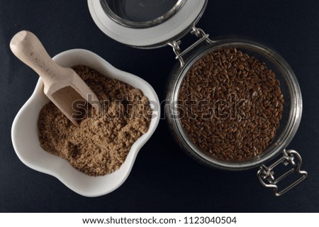 whole brown flax seeds in a jar and ground flax seeds, or linseeds, in a bowl, on a black background, top view