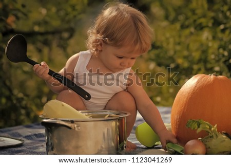 autumn harvest. Closeup of one small cook boy at picnic sitting with black ladle pot orange pumpkin and other vegetables playing with food sitting on blue checkered plaid on natural background