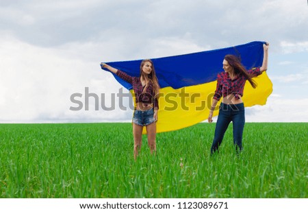 two young beautiful Ukrainian girl with an excellent figure unfurled the Ukrainian yellow and blue flag against the blue sky and the green g wheat field  