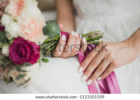 Beautiful colorful wedding flowers in hands of young bride sitting alone in home interior. Fingernails with beautiful bridal manicure and engagement ring. Horizontal color photography.
