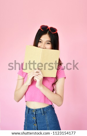 beautiful portrait Asian girl with red heart shaped sunglasses holding notebook and smart phone, cute woman in casual clothes with beaming smile standing on pink background