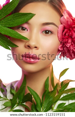 Close up isolated portrait of a young Asian lady accessorized her hairdo with real peony flowers. The beautiful girl wearing pink eye shadow and lip gloss, looking at the camera through green leaves.