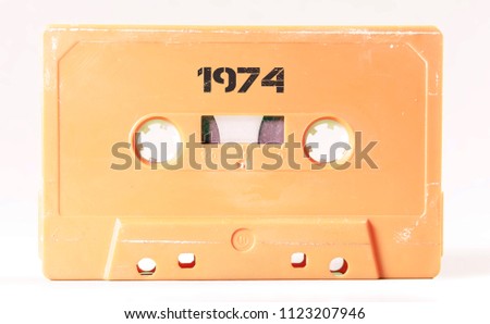 A vintage cassette tape from the 1980s era (obsolete music technology) with the text 1974 printed over it, stencil font. Color: cream, sand. White background.