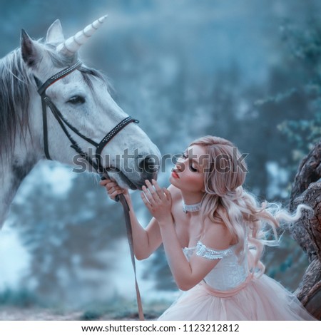A fairy in a tender vintage dress hugs a unicorn. Fantastic magical, radiant horse. Background river and forest. Blonde girl with wavy hair - light elf. Artistic Photo
