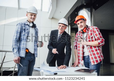The builders and businessman are discussing the construction plan in the modern business center. A man in a classic costume shows in the distance. working form in protective helmets.