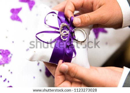 A young boy carefully handles some wedding rings on a white cushion with a purple ribbon in a bow