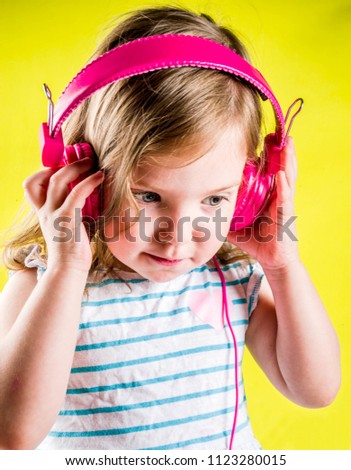 Funny cute blonde toddler girl listen music with bright pink headphones, yellow background copy space