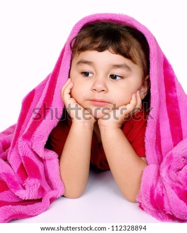 Cute four-year-old girl with nothing to do looks out pensively from under a velvety soft pink blanket, isolated on white background