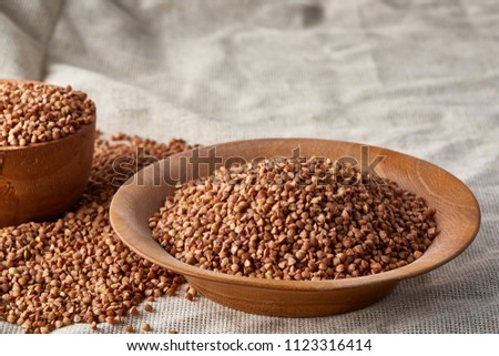 Bowl and plate full of buckwheat grains on homespun tablecloth, close-up, selective focus, shallow depth of field.