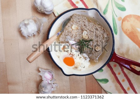 Spanish Castilian garlic soup with bread, egg and rosemary (Sopa de Ajo Castellana)(enameled cast iron, wooden spoon, wooden table, pink garlic cloves)