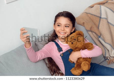 Little girl at home sitting on sofa hugging teddy bear taking selfie photos on smartphone smiling happy