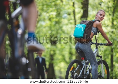 Focus on sporty lady exploiting bicycle in green environment. She is turning around to look at person riding behind her. Female is grinning and carrying backpack