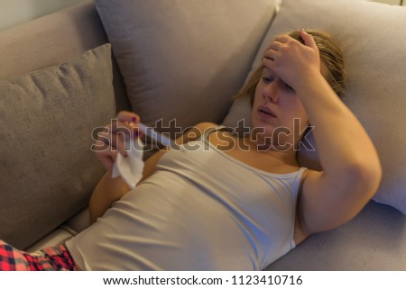 Sick woman lying in bed with high fever. Cold flu and migraine. Sick young woman at home on the sofa, she is covering with a blanket, taking temperature 