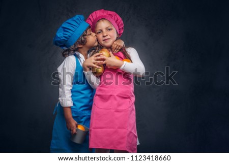 Cute cook couple. Little boy with brown curly hair dressed in a blue cook uniform and beautiful schoolgirl dressed in a pink cook uniform, poses with cookware and fruits at a studio. Isolated on the